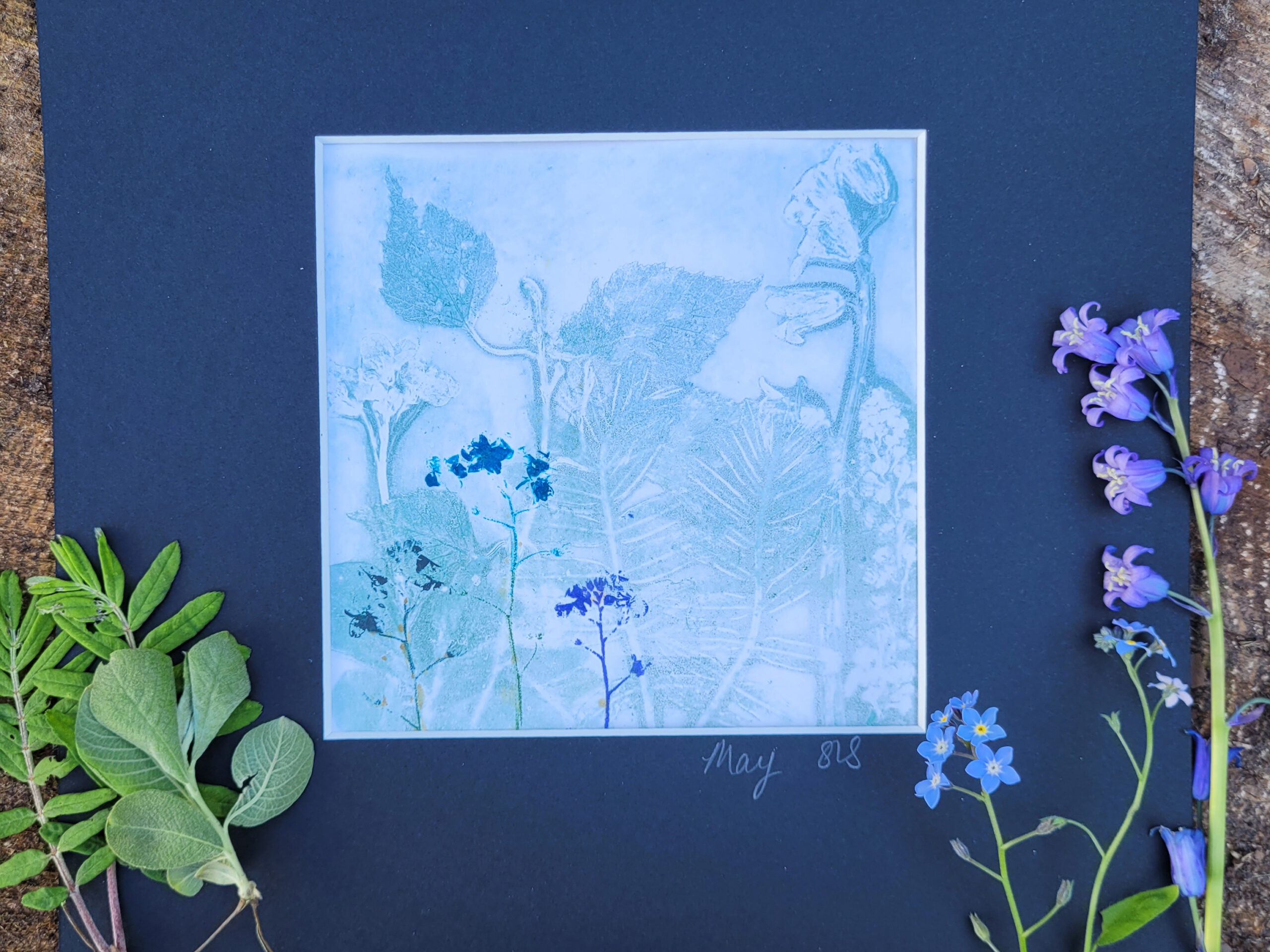 A blue press printing of leaves and flowers on a blue mount. Green leaves and blue flowers rest on top of the mount.