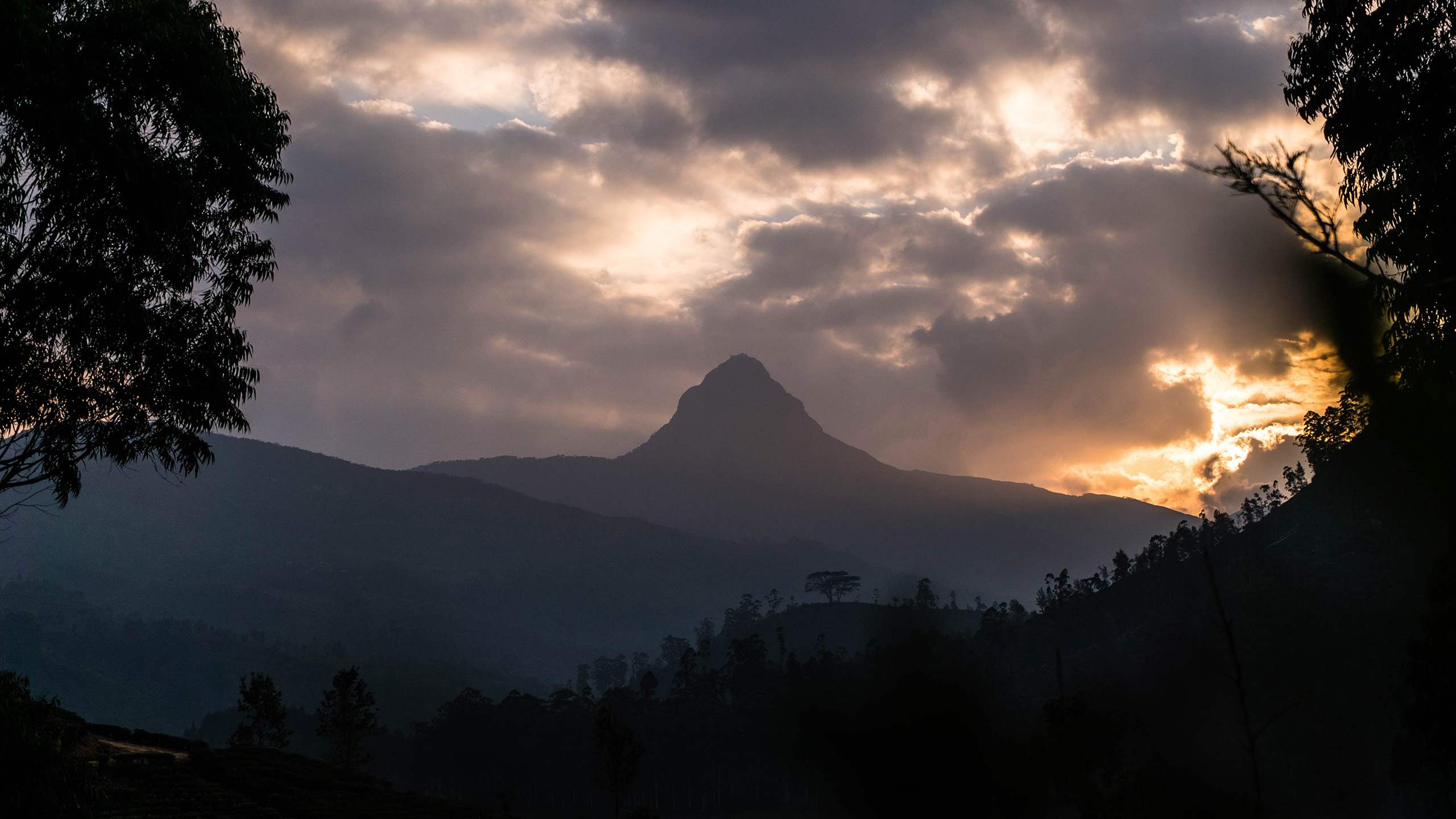 A mountain in the distance with the sun peaking through grey clouds.