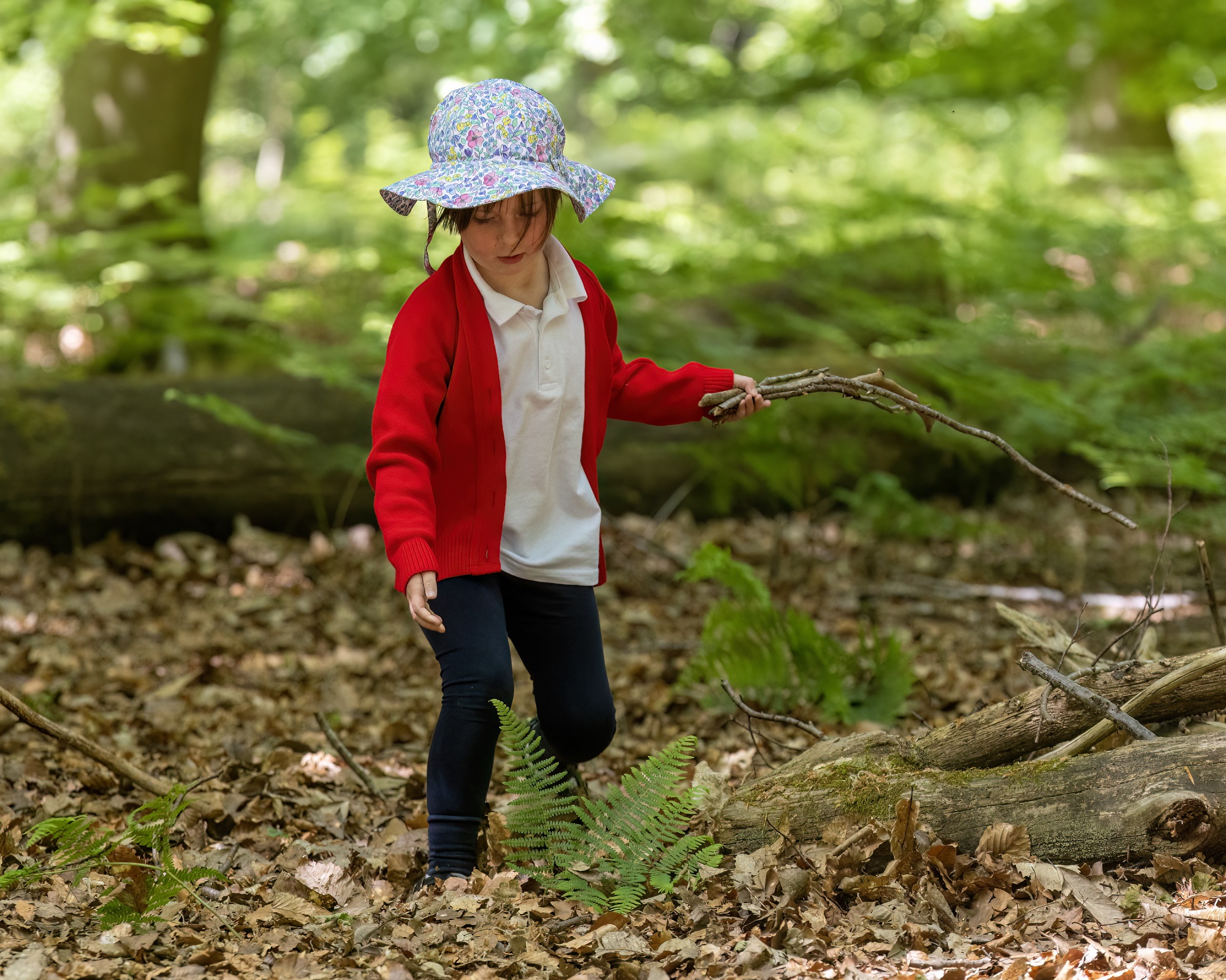 Young Child wearing a floral hat and red cardigan holding sticks.