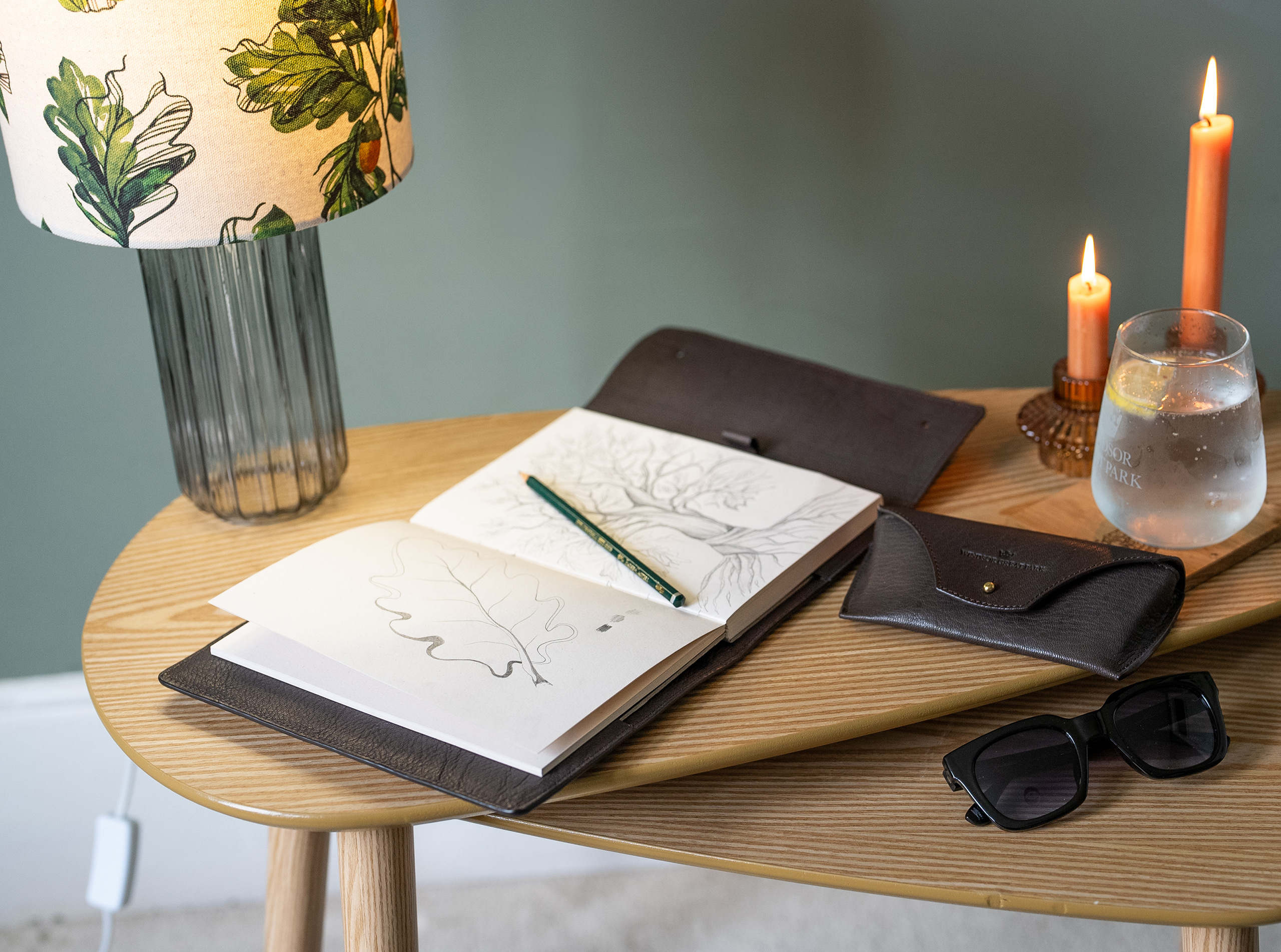 Wooden tables with a lamp, candles, leather journal and glasses case with glasses.