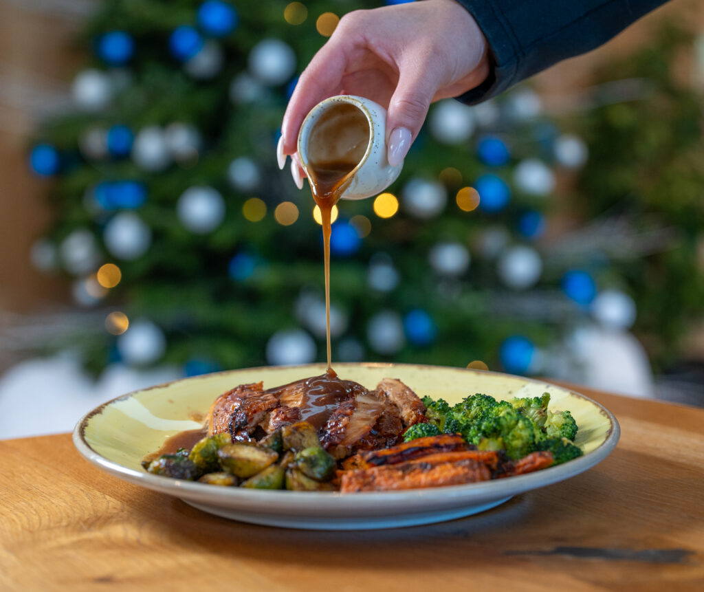 Pouring gravy on to a roast lunch with a Christmas Tree in the background.